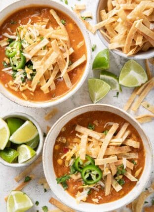 Two bowls of tortilla soup garnished with homemade tortilla strips. A bowl of tortilla strips and a bowl of lime wedges sits next to the soup bowls.