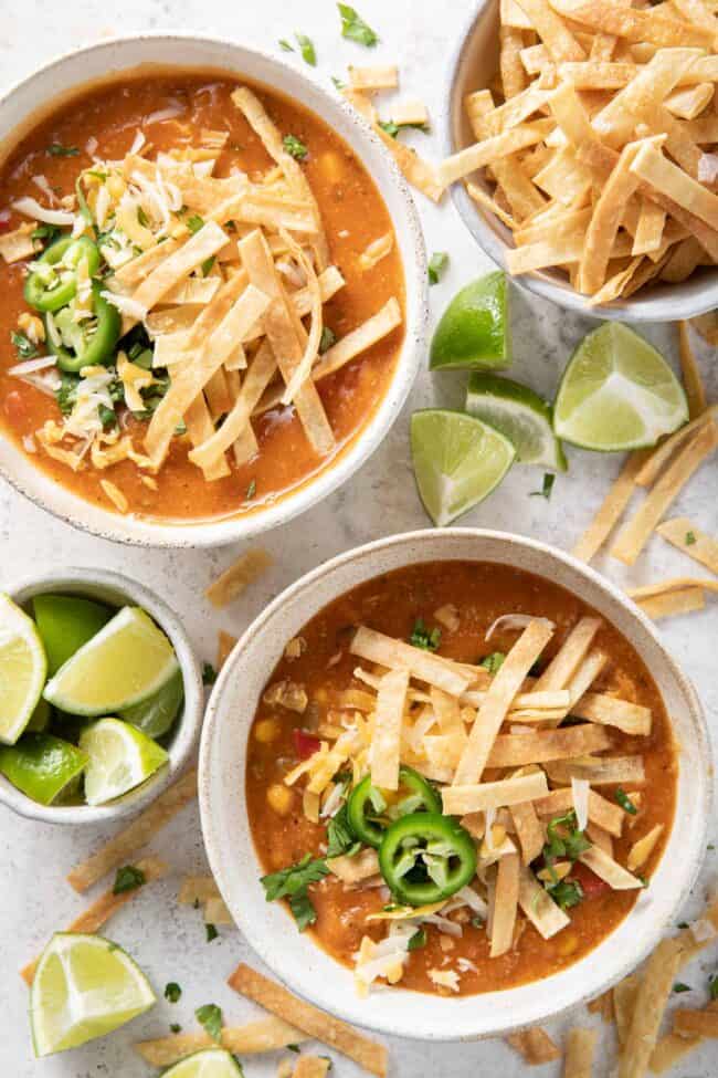 Two bowls of soup topped with homemade tortilla strips. A bowl of tortilla strips sits next to the soup bowls.