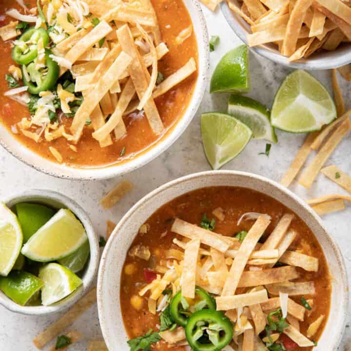 Two bowls of tortilla soup garnished with homemade tortilla strips. A bowl of tortilla strips and a bowl of lime wedges sits next to the soup bowls.