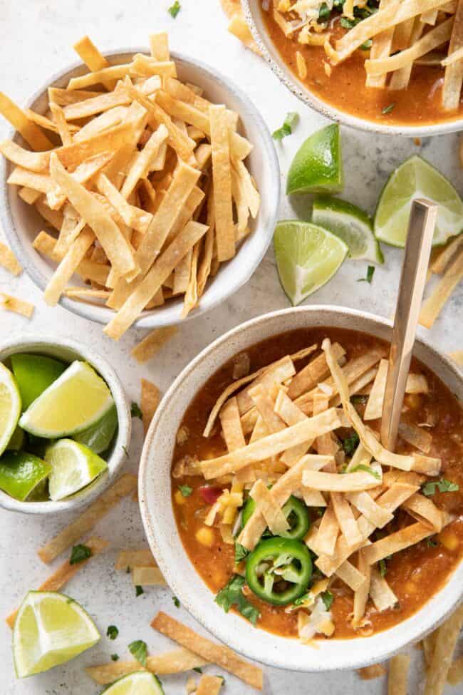A bowl of tortilla soup garnished with tortilla strips. A bowl of tortilla strips and a bowl of lime wedges sits next to the soup bowl.