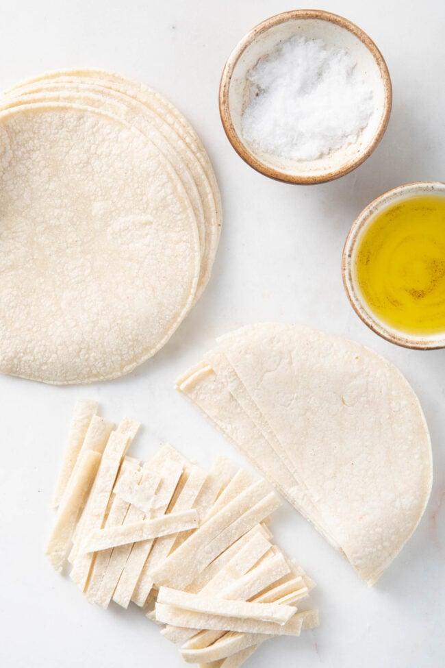 Corn tortillas on a cutting board, some are cut into thin strips. A small bowl of olive oil and a bowl with salt sit next to the tortillas.