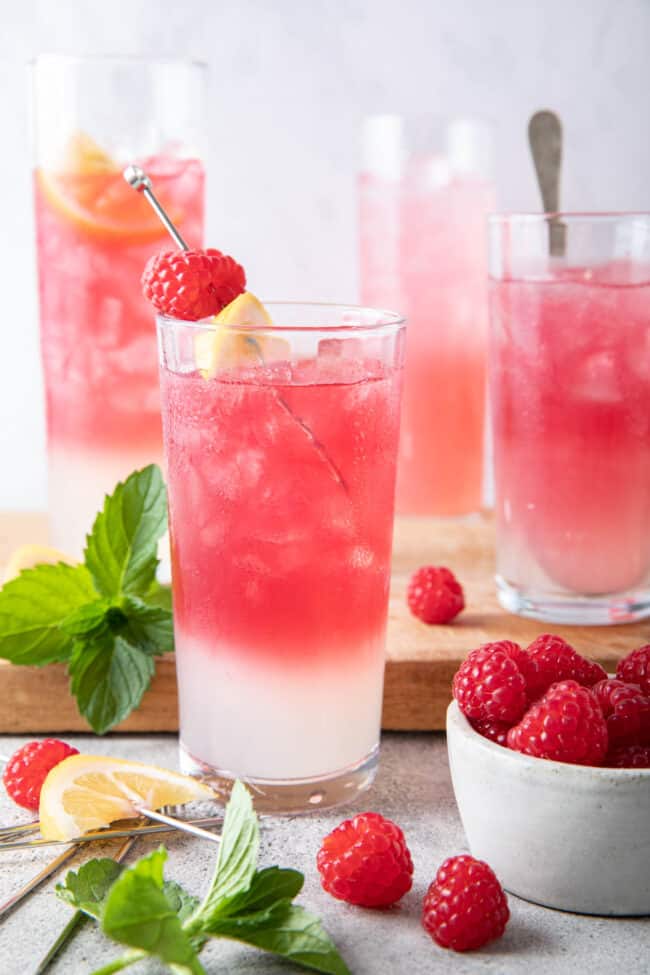 Four dinking glasses filled with rose colored drinks. A small bowl filled with raspberries sits next to the glasses.