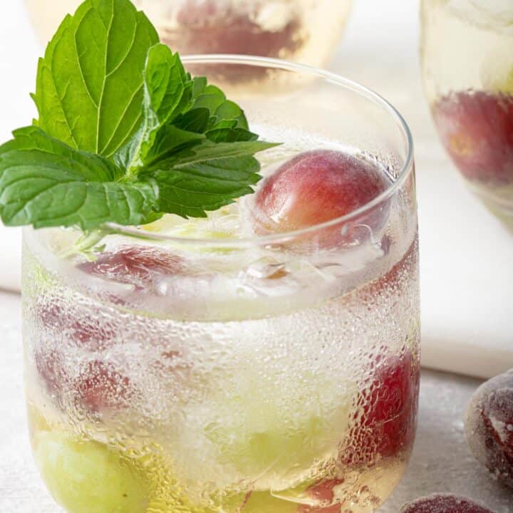 A clear glass filled with white grape spritzer and frozen grapes. A sprig of mint garnishes the glass.