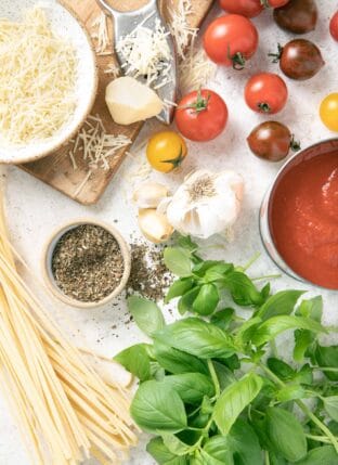 A bunch of basil leaves sit next to a can of tomato sauce, dried noodles, garlic and parmesan cheese for basil substitute.