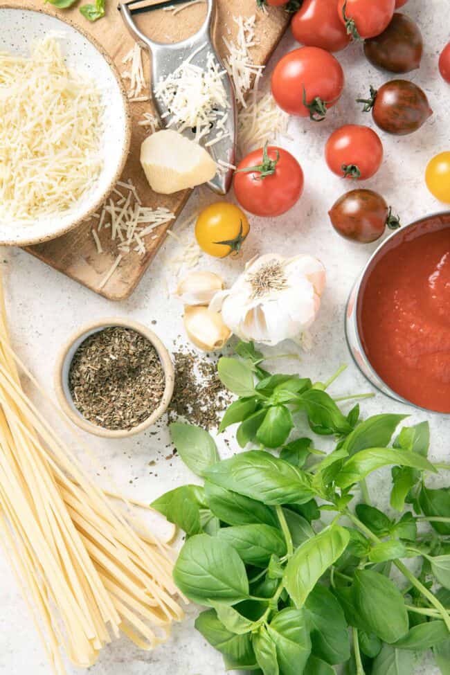 Fresh herbs, a can of tomato paste, parmesan cheese and cherry tomatoes sit on a wooden board. 