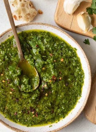 A white bowl filled with chimichurri sauce. A gold spoon sits in the bowl.