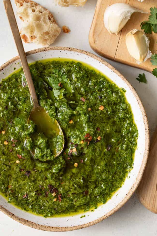 A white bowl filled with chimichurri sauce. A gold spoon sits in the bowl.