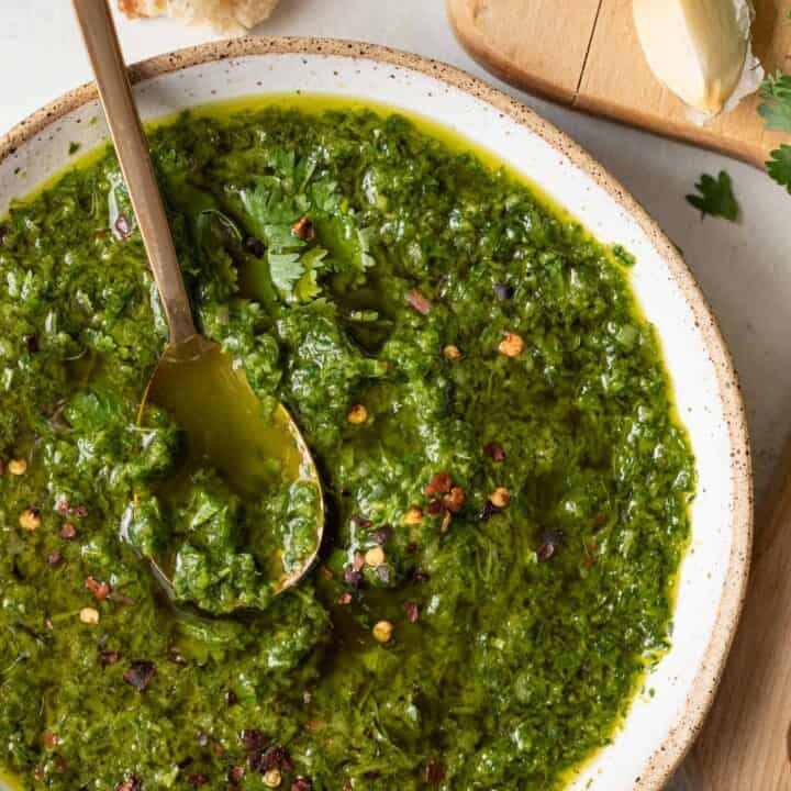 A white bowl filled with chimichurri sauce. A gold spoon sits in the bowl.