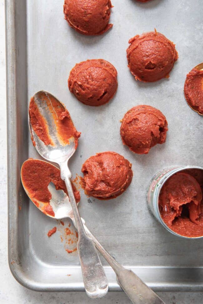 Two tablespoons sit next to a can of tomato paste and several scoops of tomato paste on a cookie sheet.