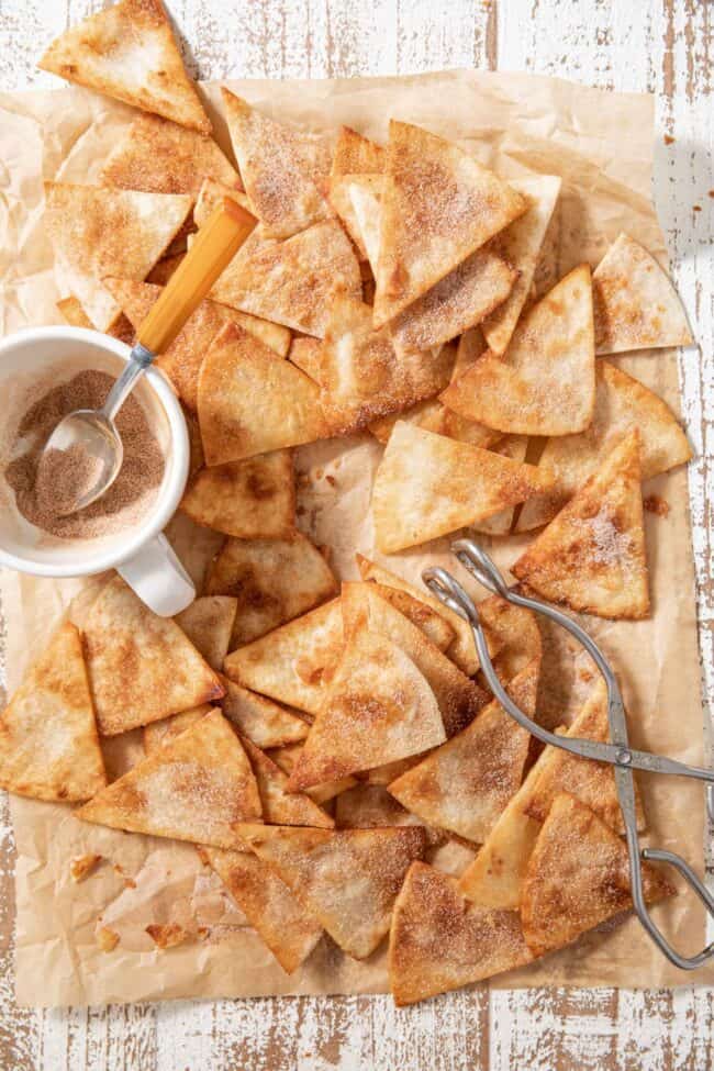 A lot of fried tortilla chips (churro chips) dusted in cinnamon and sugar are laying on parchment paper. A cup with cinnamon sugar sits next to the chips.