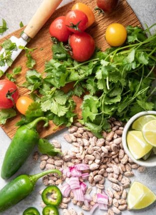 A brown cutting board with a knife sitting on it next to sprigs of fresh cilantro, tomatoes and jalapeno.