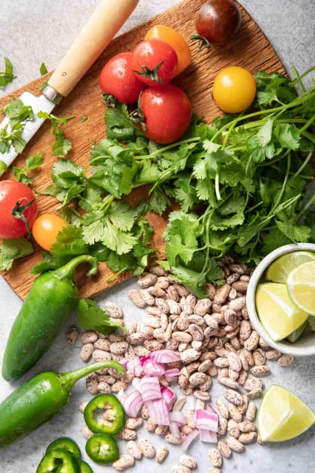 A brown cutting board with a knife sitting on it next to sprigs of fresh cilantro, tomatoes and jalapeno.