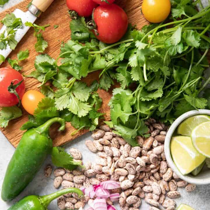 A brown cutting board with a knife sitting on it next to sprigs of fresh cilantro, tomatoes and jalapeno.