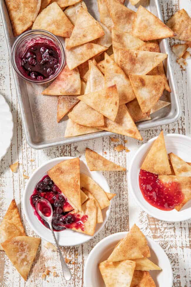 A cookie sheet filled with fried cinnamon tortilla chips (churro chips). Two white plates with the chips on them sits next to the cookie sheet.