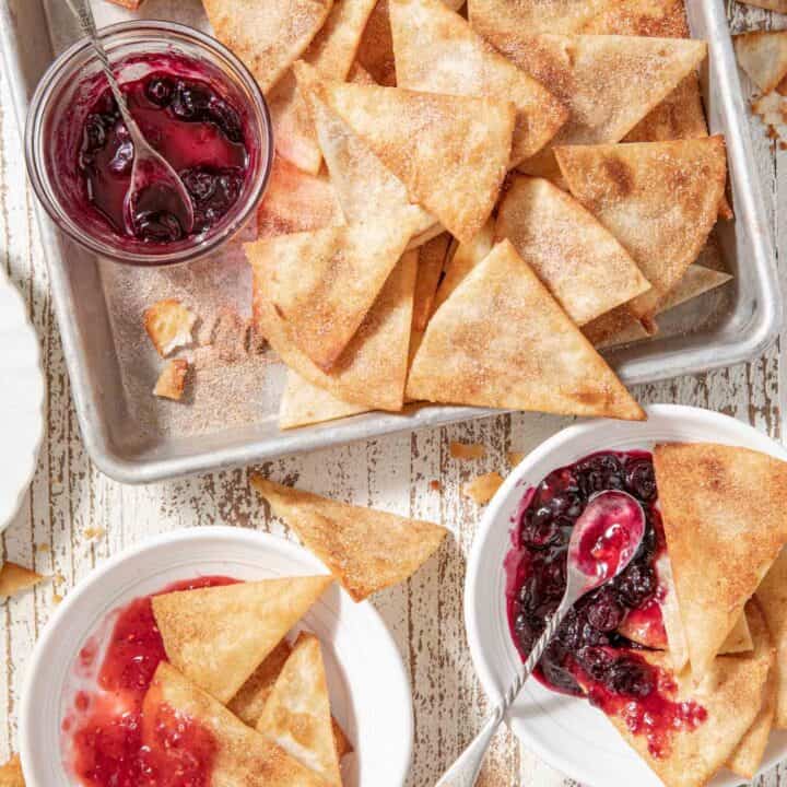 A cookie sheet filled with fried cinnamon tortilla chips. Two white plates with the chips on them sits next to the cookie sheet.