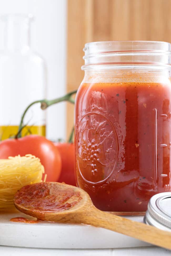 A clear glass mason jar filled with marinara sauce. A wooden spoon rests next to the jar.