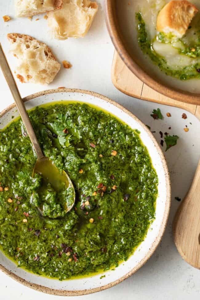 A white bowl filled with chimichurri sauce. A bowl of soup with chimichurri swirled in it sits next to the sauce bowl.