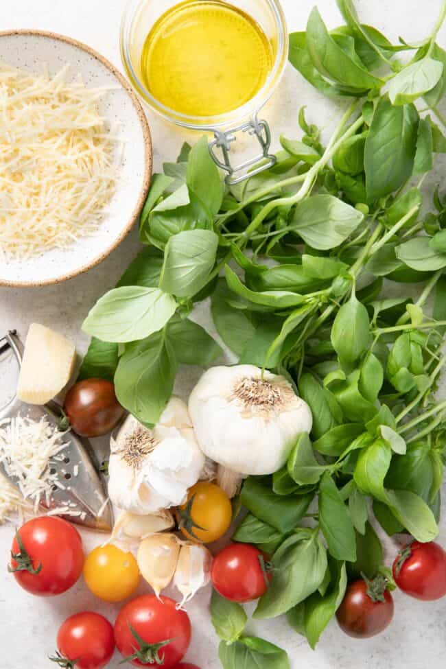 A bunch of basil leaves sit next to garlic cloves, a small bowl of parmesan cheese and a small jar of olive oil for substitute for basil.