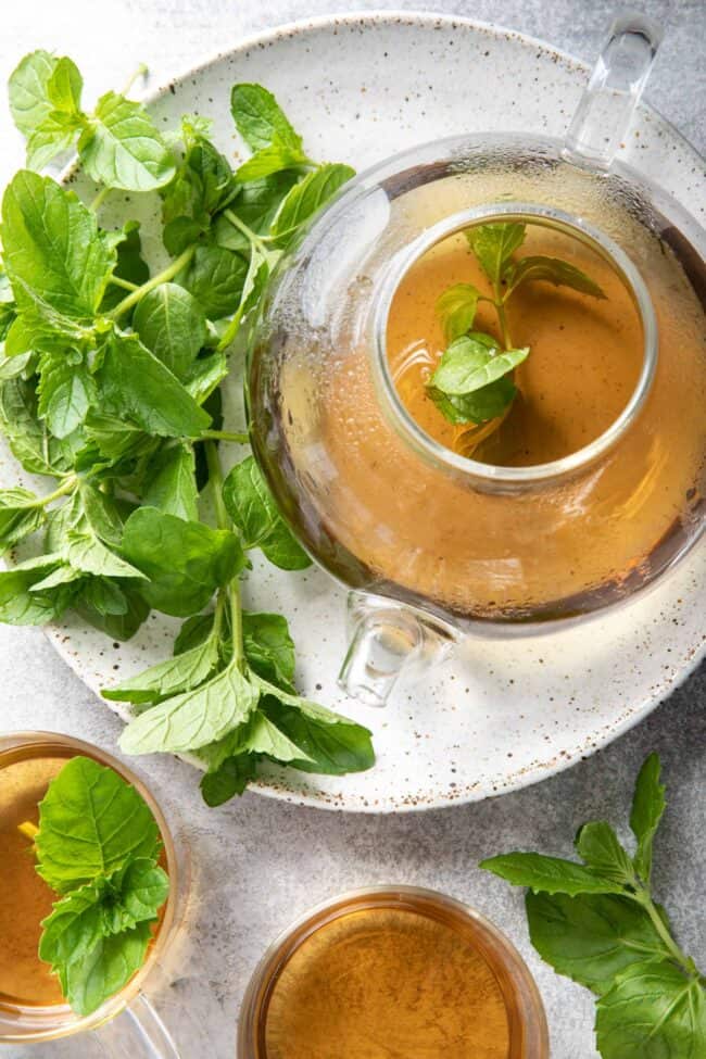 A clear glass tea pot filled with tea sits on a white plate next to a bunch of mint leaves.