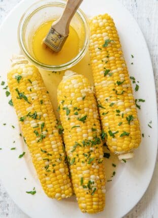 A white platter with 3 ears of boiled corn on it brushed with butter and dusted with chopped cilantro.