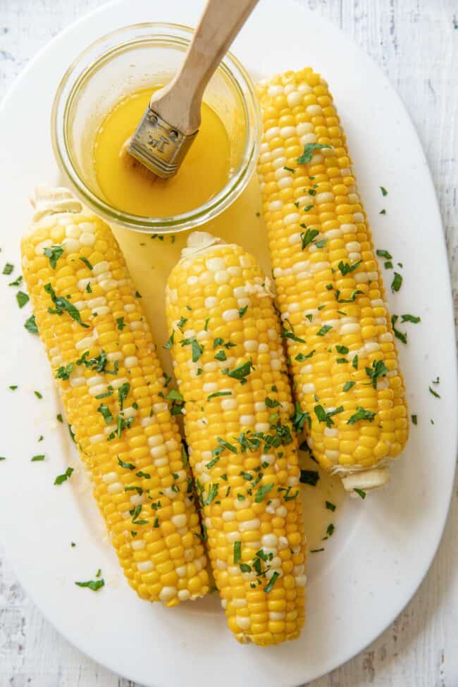 Three corn cobs of boiled corn on a white platter. A glass bowl with melted butter sits next to the corn.