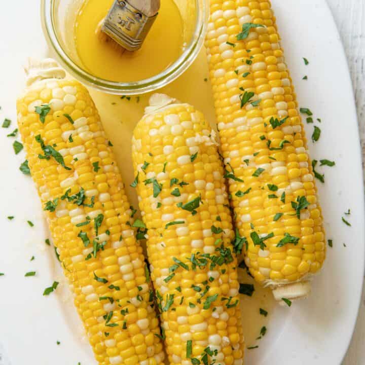 A white platter with 3 ears of boiled corn on it brushed with butter and dusted with chopped cilantro.