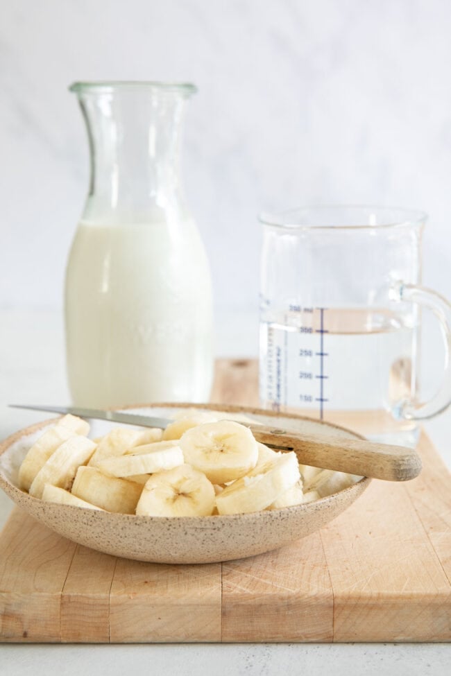 A bowl of sliced bananas with a knife resting on the bowl. A bottle of milk and a measuring cup filled with water sits next to the bowl.