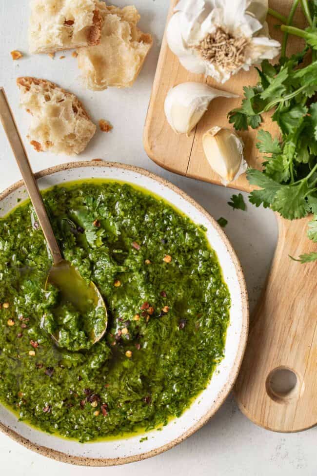 A white bowl filled with green sauce. Torn pieces of crusty bread sits next to the bowl.