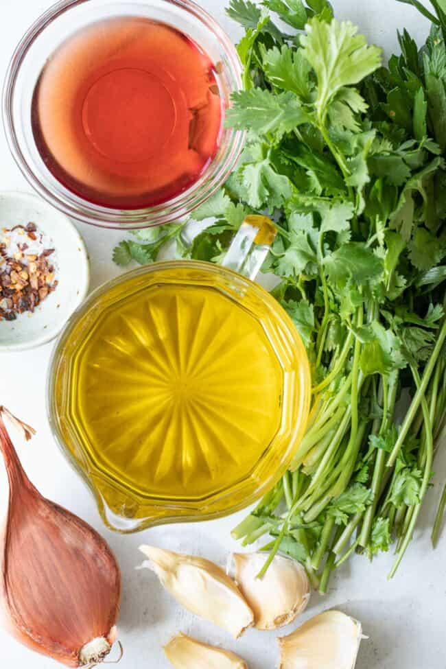 Glass measuring cups - one filled with olive oil and the other filled with red wine vinegar. Sprigs of parsley and cilantro, shallot and cloves of garlic sit next to the measuring cups.