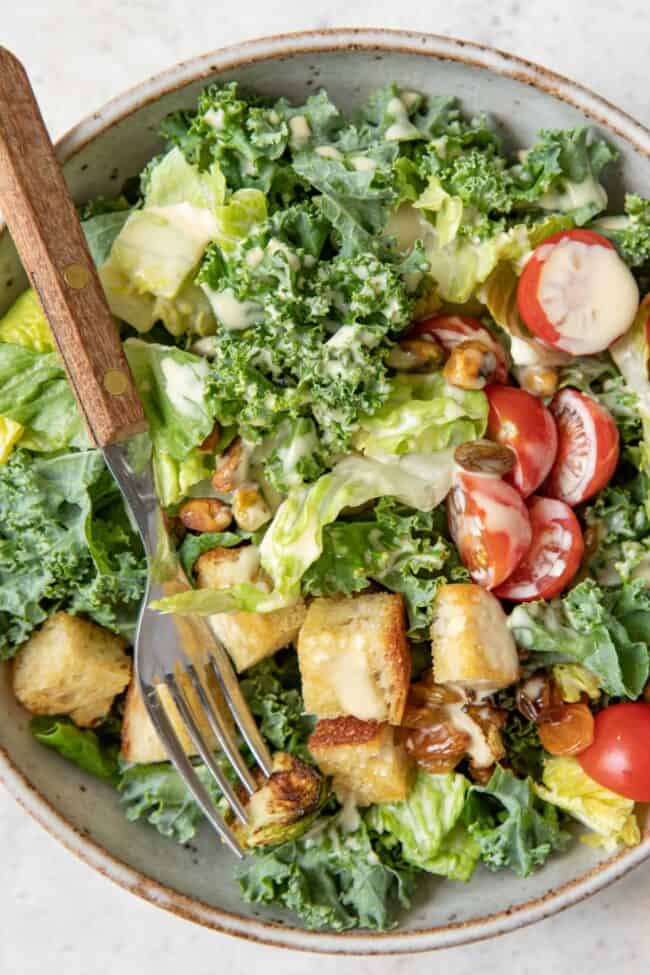 A bowl filled with Kale Caesar salad (tossed with Romaine lettuce, chopped kale, croutons, cherry tomatoes and golden raisins).