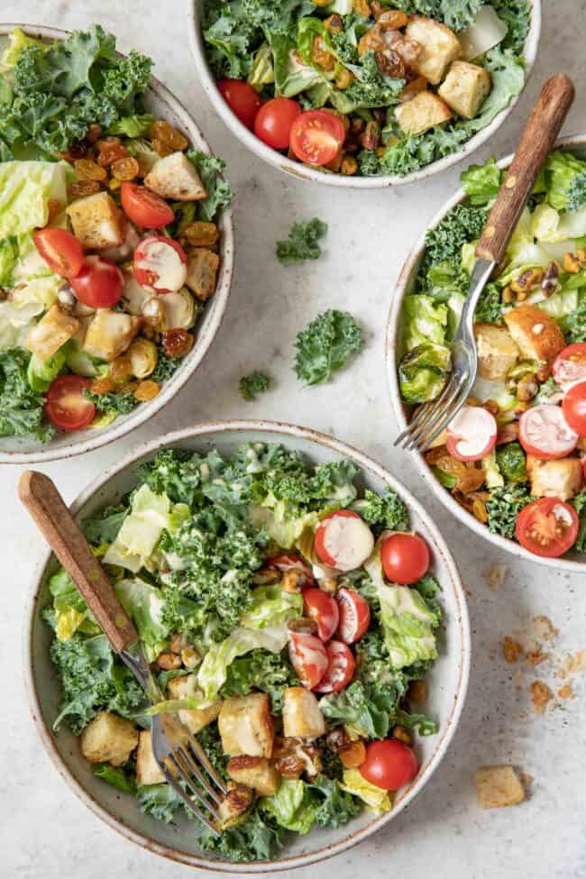Four bowls filled with Kale Caesar Salad (chopped kale, Romaine lettuce, croutons, cherry tomatoes). Two forks with brown wooden handles are poked in two bowls.
