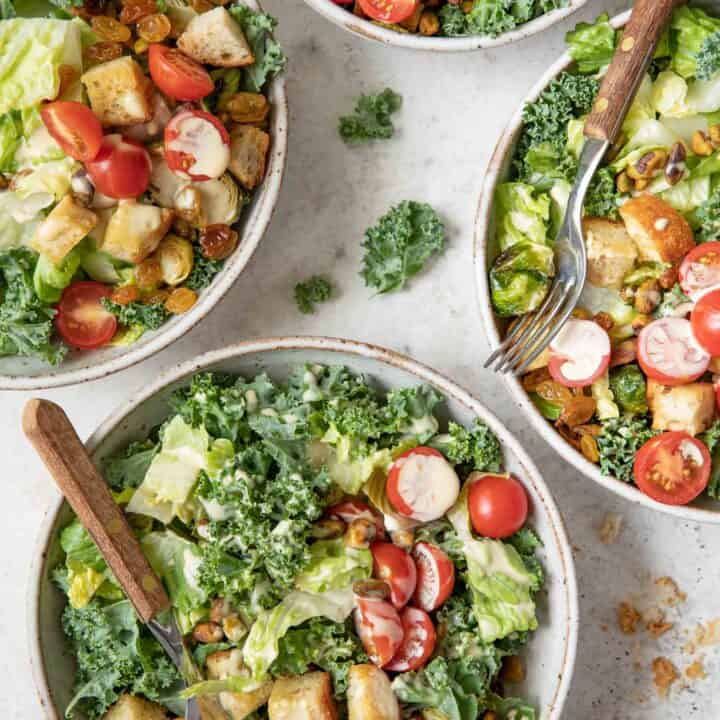 Four bowls filled with Kale Caesar Salad (chopped kale, Romaine lettuce, croutons, cherry tomatoes). Two forks with brown wooden handles are poked in two bowls.