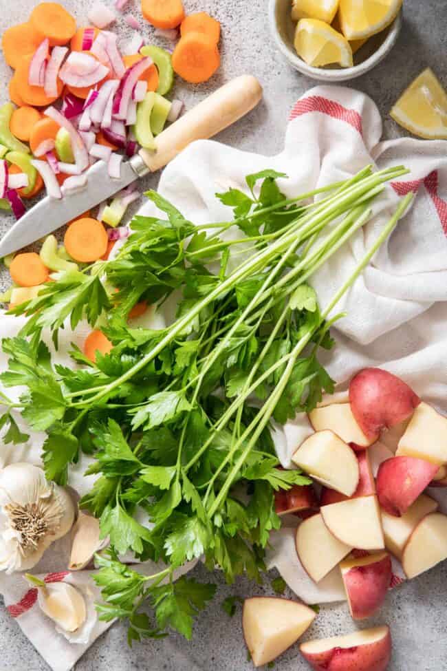 Fresh vegetables (red potatoes, carrot, celery and onion) are chopped next to a kitchen towel and kitchen knife. 