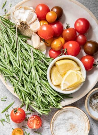 A white plate with a bunch of rosemary sprigs on it next to garlic bulbs and cherry tomatoes.