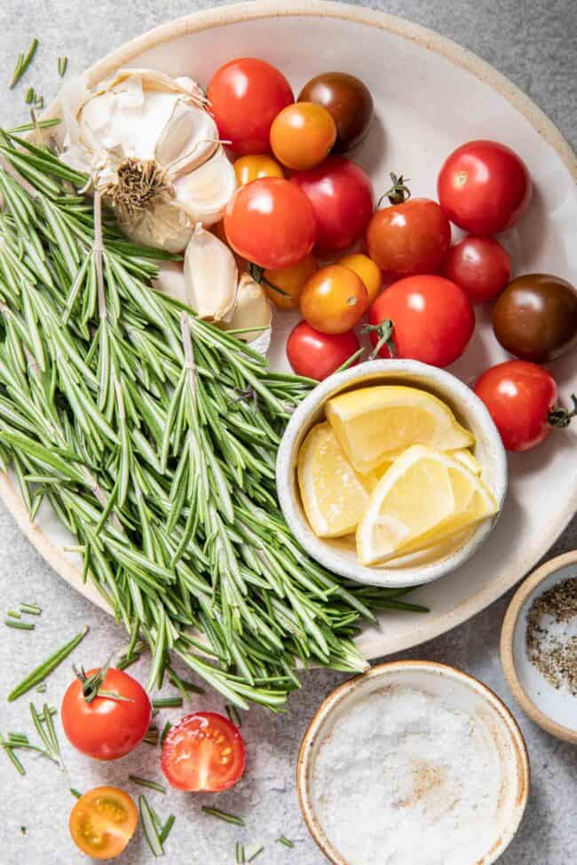 A white plate with a bunch of rosemary sprigs on it next to garlic bulbs and cherry tomatoes.