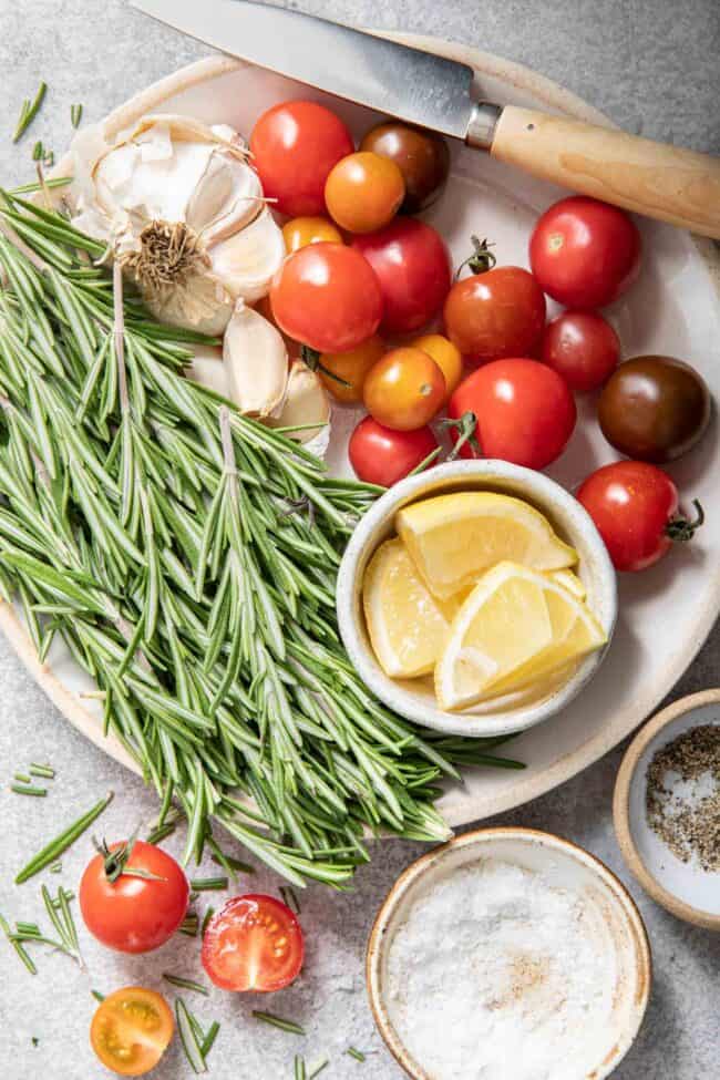 A white plate with a bunch of rosemary sprigs on it next to garlic bulbs and cherry tomatoes.