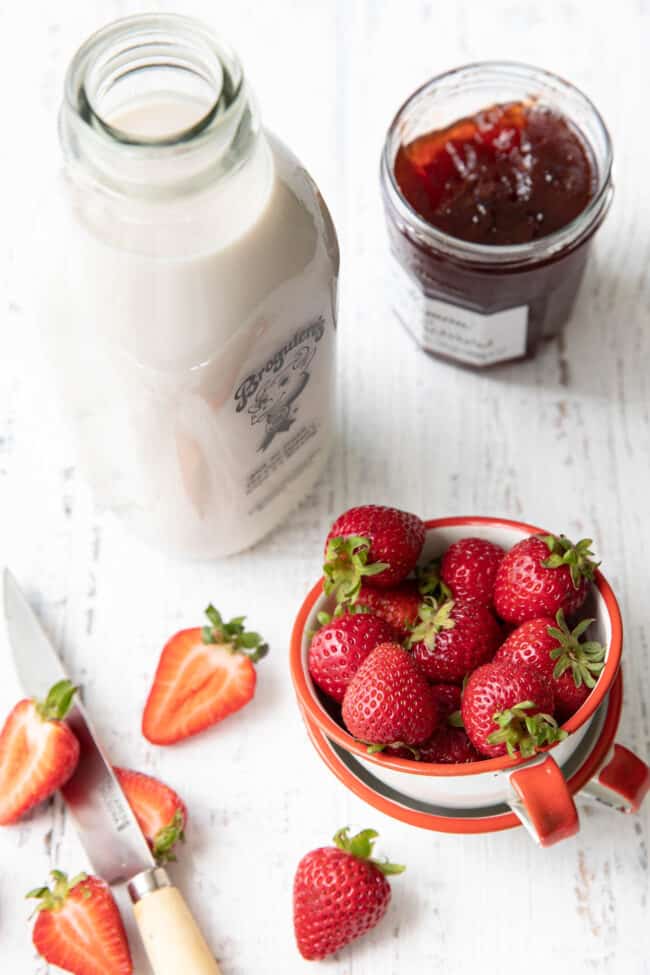 Fresh strawberries in a white cup sits next to a knife with strawberries sliced in half. A bottle of milk and jar of preserves sit next to the strawberries.