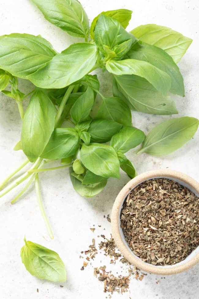 A bunch of fresh basil leaves sit next to a small bowl of dried basil.