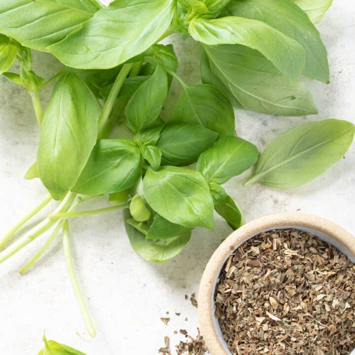 A bunch of basil leaves sit next to a small bowl filled with dried basil.