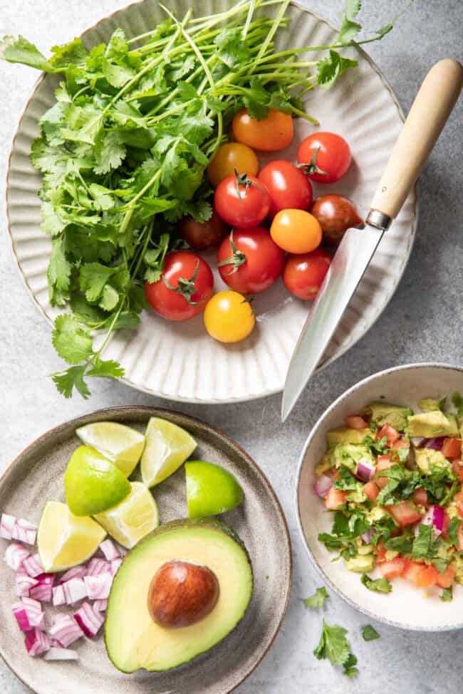 A gray plate with a bunch of cilantro sprigs sit next to cherry tomatoes. A knife rests on the plate. A smaller plate has half of an avocado with lime wedges and chopped parsley sits next to it.