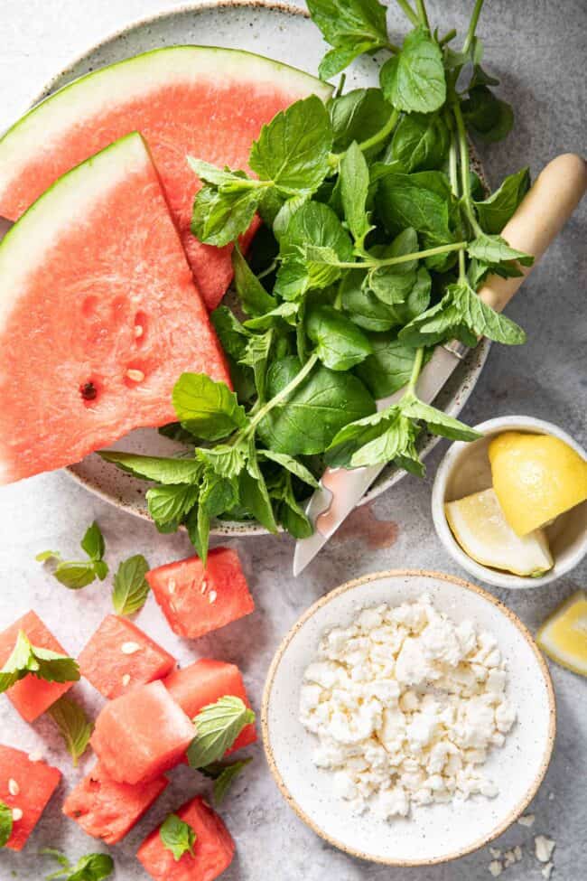 Slices of watermelon and a bunch of fresh herbs sit on a white plate together.