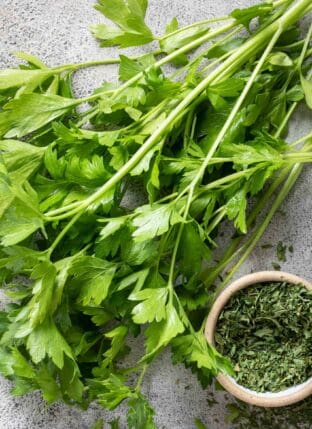 A small bunch of fresh parsley lays next to a small bowl filled with dried parsley (for parsley substitute).