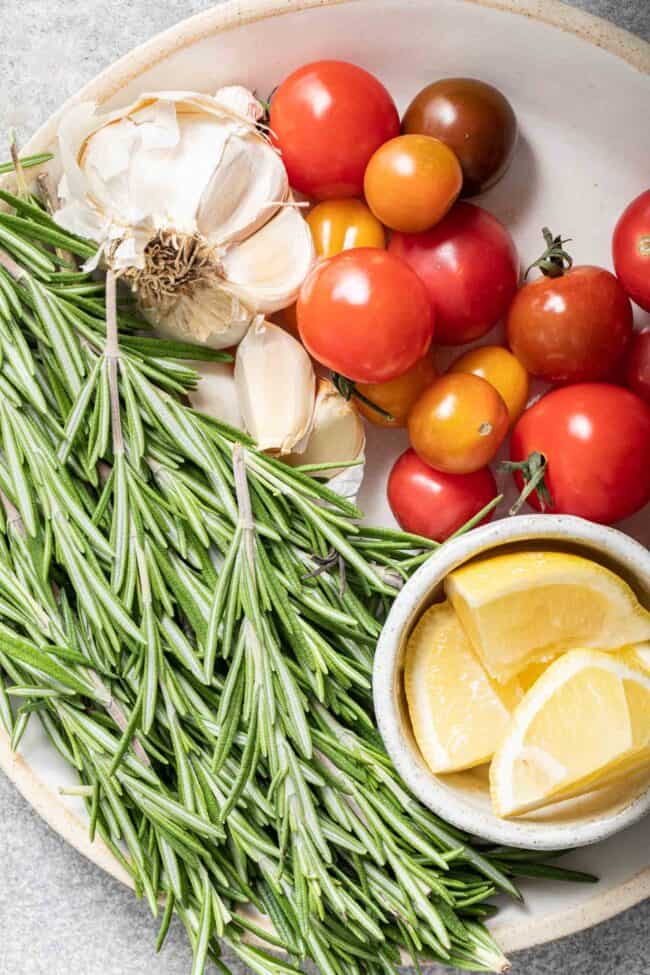 A white plate with a bunch of rosemary sprigs on it next to garlic bulbs and cherry tomatoes.