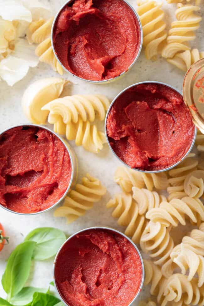Four open cans of tomato paste. Curly pasta noodles are scattered around the cans. For tomato paste substitute.