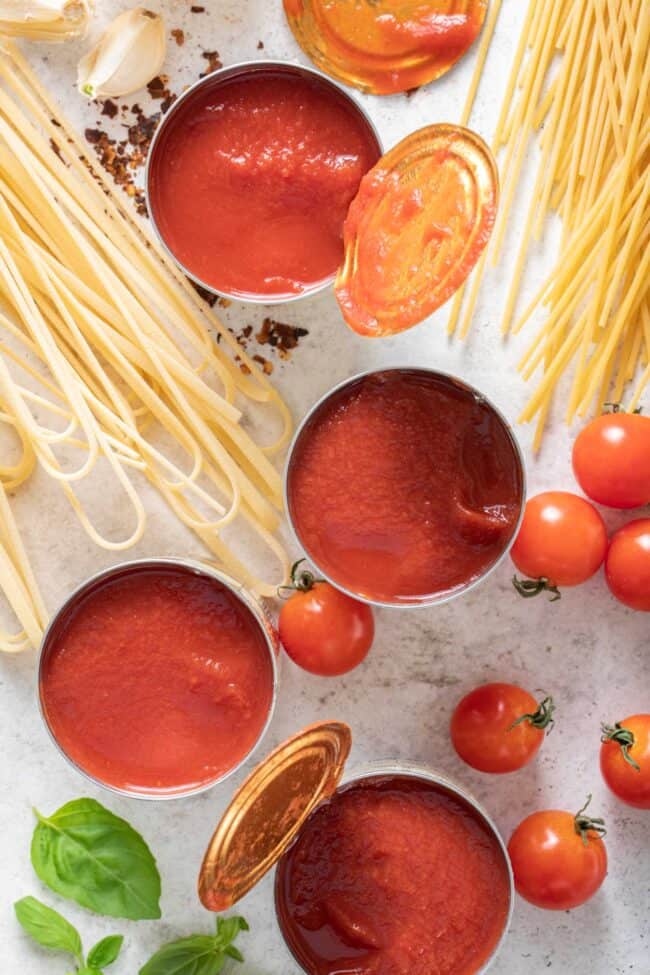 Four cans of opened tomato sauce sitting next to uncooked pasta noodles, basil and tomatoes.