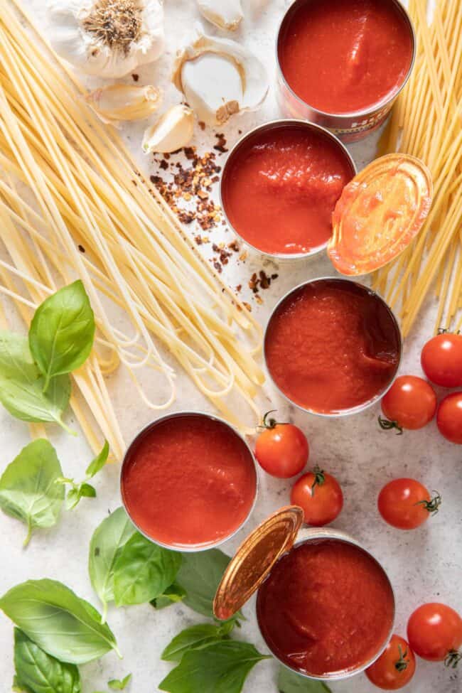 Five cans of opened tomato sauce next to uncooked noodles, fresh basil and tomatoes.