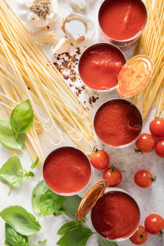 Five cans of opened tomato sauce next to uncooked noodles, fresh basil and tomatoes.