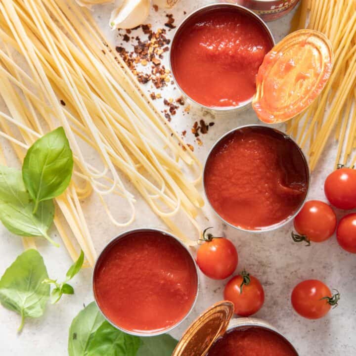 Five cans of open tomato sauce next to uncooked noodles, basil leaves and cherry tomatoes.