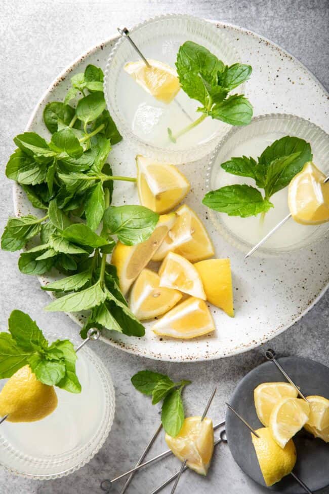 Two clear glasses filled with lemonade sits on a white plate with fresh basil sitting next to it. 