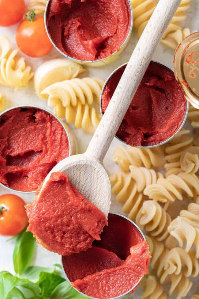 Four open cans of tomato paste. A wooden spoon with a heaping scoop of paste rests on two of the cans.